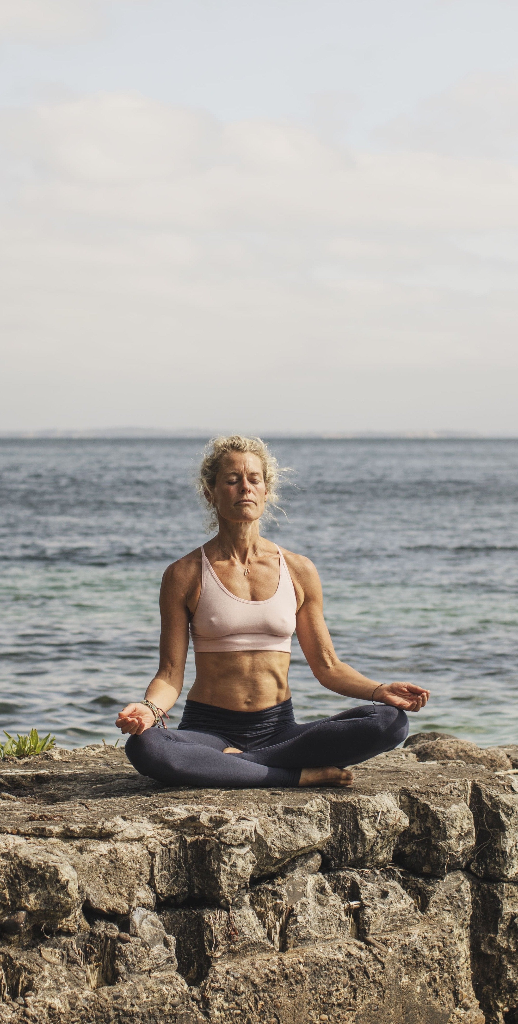 Woman sitting cross-legged in a meditation pose on a rock by the sea, eyes closed.